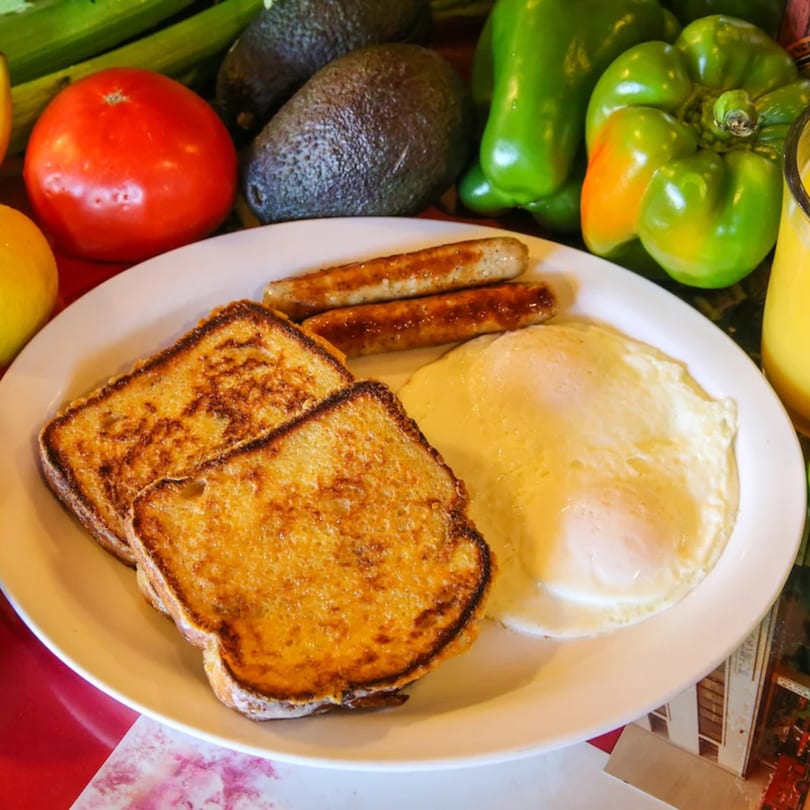 Breakfast plate with French toast, sausage links, and fried eggs, surrounded by fresh produce.