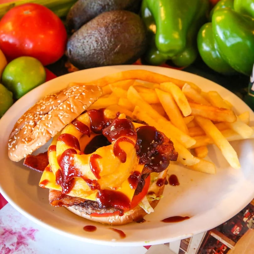 A burger with cheese, bacon, pineapple, and BBQ sauce, served with fries on a white plate.