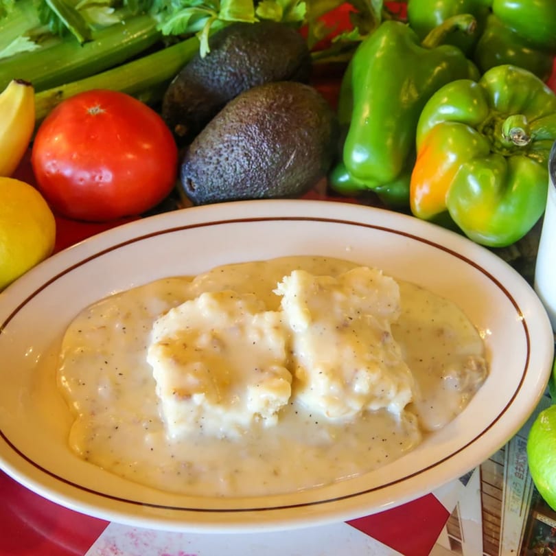 Biscuits and gravy on a white plate, surrounded by fresh vegetables like bell peppers, avocados, and tomatoes.